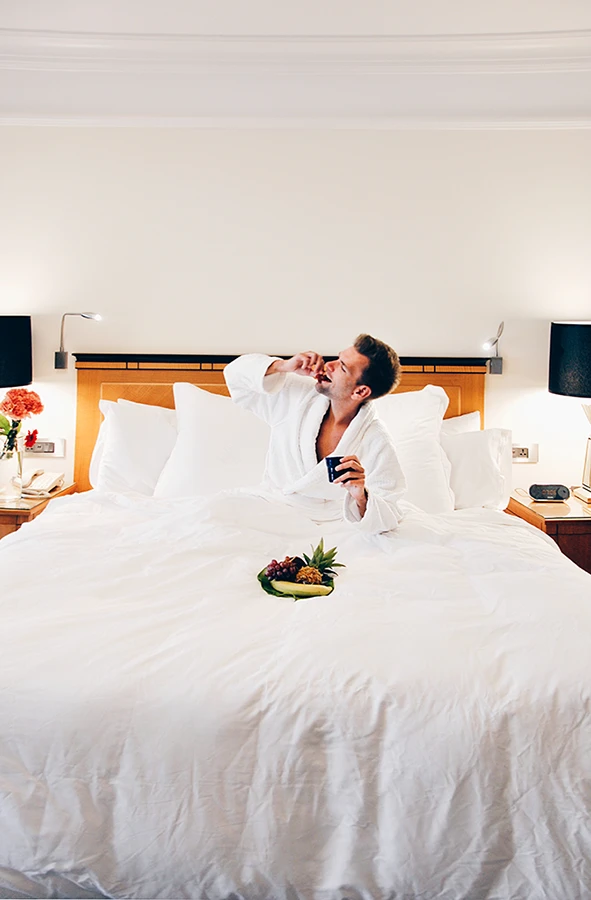 Ein Mann, der im Bademantel und einem Obstteller im Bett seines Hotelzimmers sitzt und eine Weintraube isst.