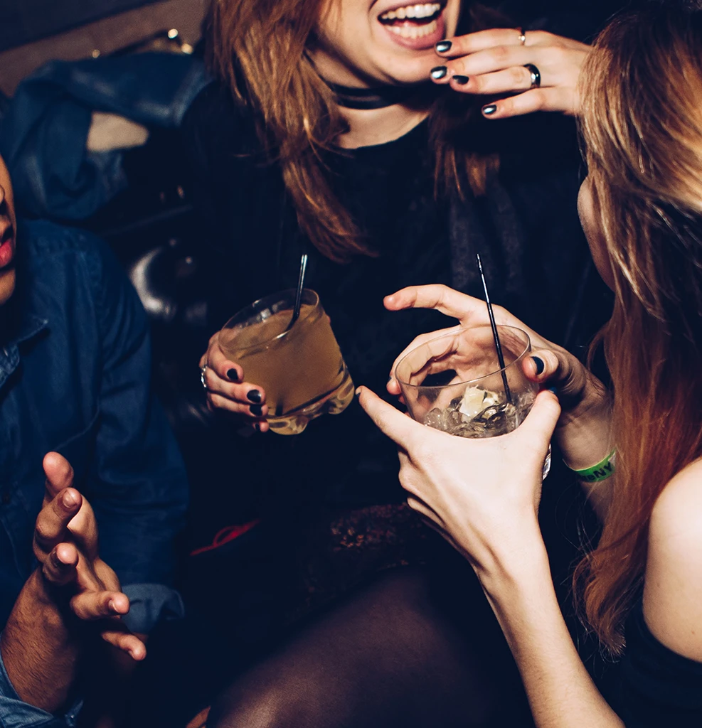 Drei Personen in einer Bar, die miteinander sprechen und lachen, während sie Getränke in ihren Händen halten.