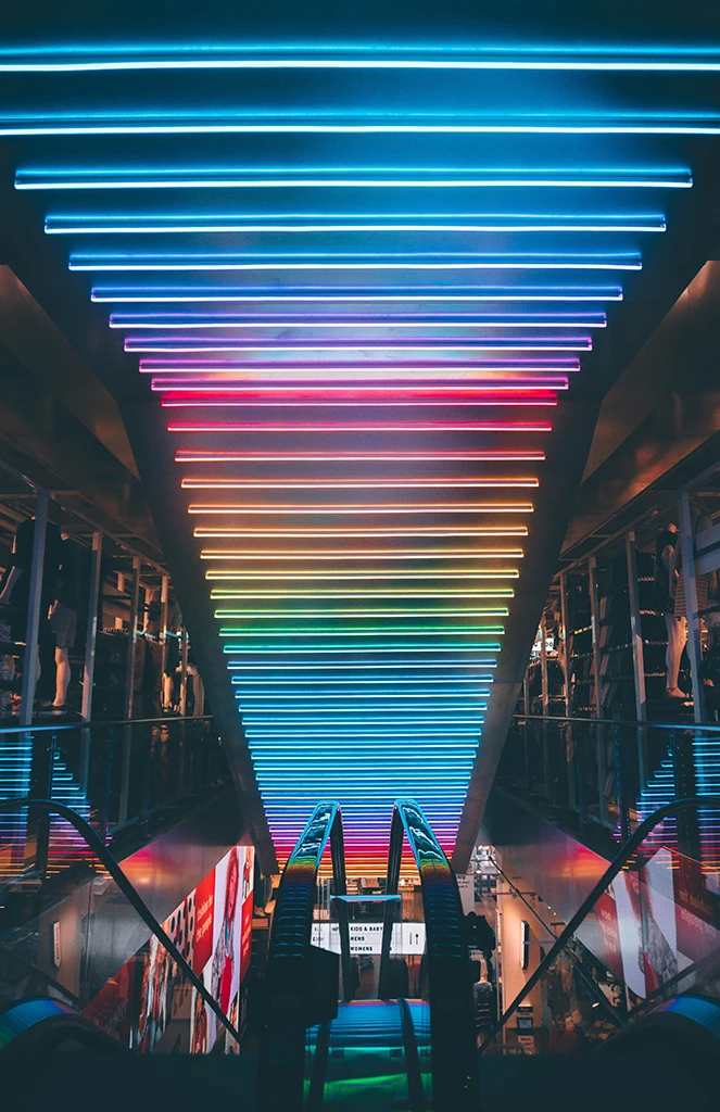 Rolltreppe nach unten, die in eine Einkaufspassage führt mit bunten Neonlichtern